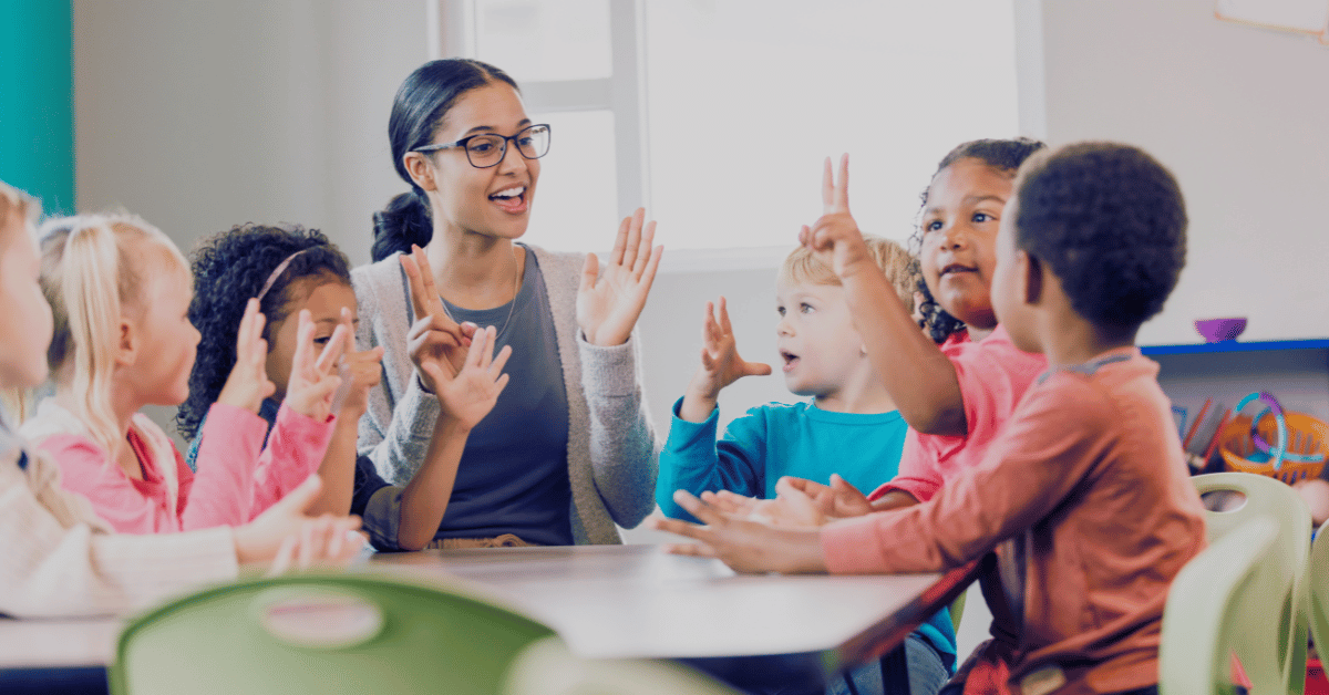 a teacher and a group of children sitting at a table with their hands up